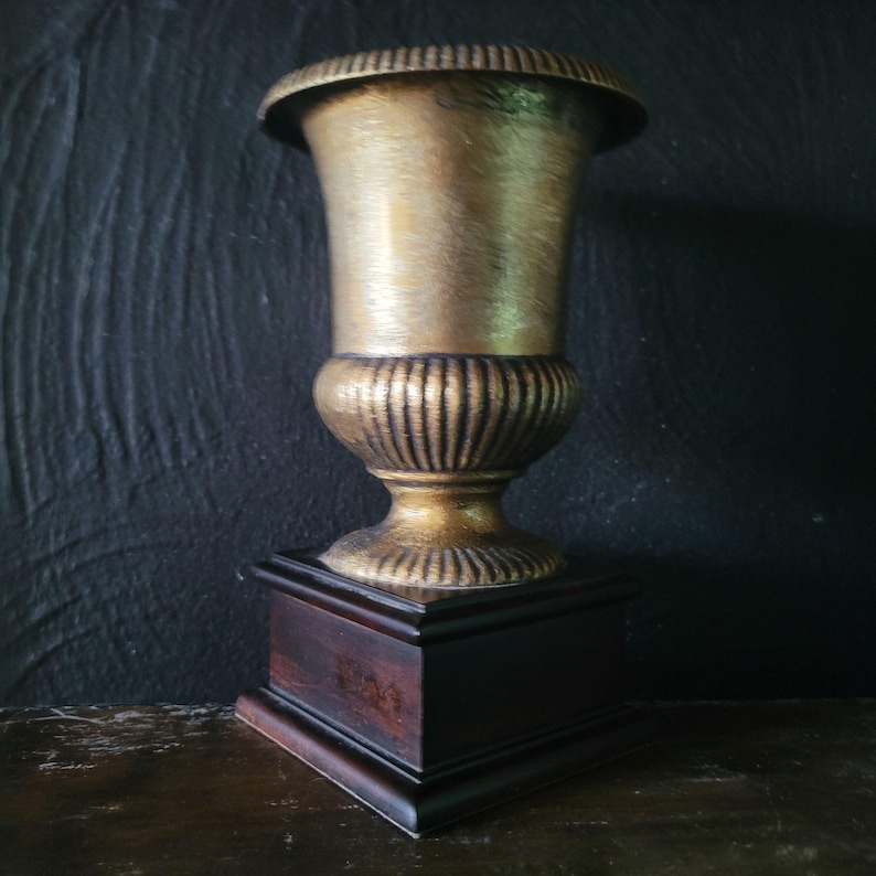 May include: A gold-colored metal vase with a fluted design sits on a dark wood base. The vase is on a wooden surface.