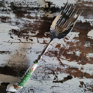 May include: A silver serving fork with a decorative handle. The handle is white with a green band and a painted Christmas tree design. The fork has a wide, slotted head and a twisted silver stem.