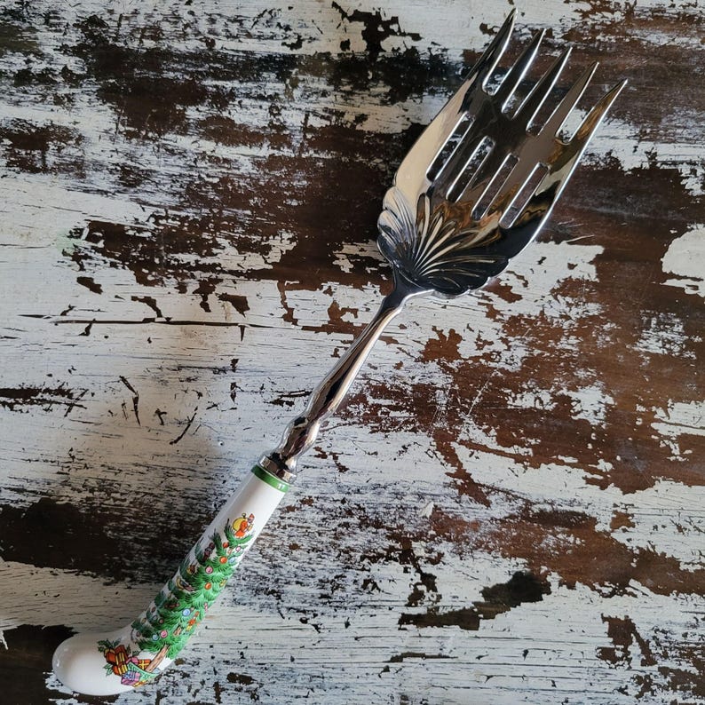 May include: A silver serving fork with a decorative handle. The handle is white with a green band and a painted Christmas tree design. The fork has a wide, slotted head and a twisted silver stem.