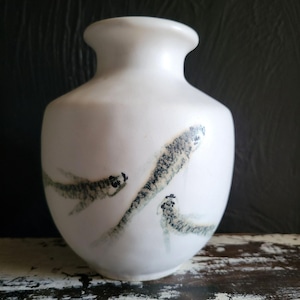 Può includere: Un vaso di ceramica bianco con corpo arrotondato e collo stretto. Il vaso è decorato con tre forme astratte a forma di pesce, grigio scuro e verde. Il vaso poggia su una superficie di legno bianco usurato.