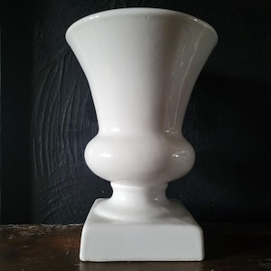 Puede incluir: Un jarrón de cerámica blanca brillante estilo urna con un borde ancho y acampanado, un cuerpo redondeado y una base cuadrada resistente. El recipiente tiene un acabado liso y reflectante con una silueta simétrica de color blanco sólido.