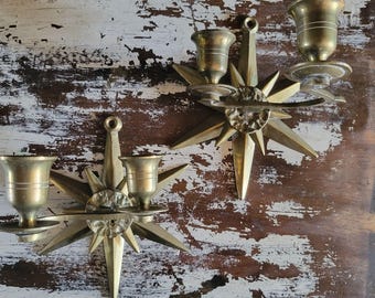 May include: Two vintage brass candle holders. Each holder features a starburst design with multiple points and a cup-shaped holder for the candle. The holders are mounted on a wooden surface with peeling white paint.