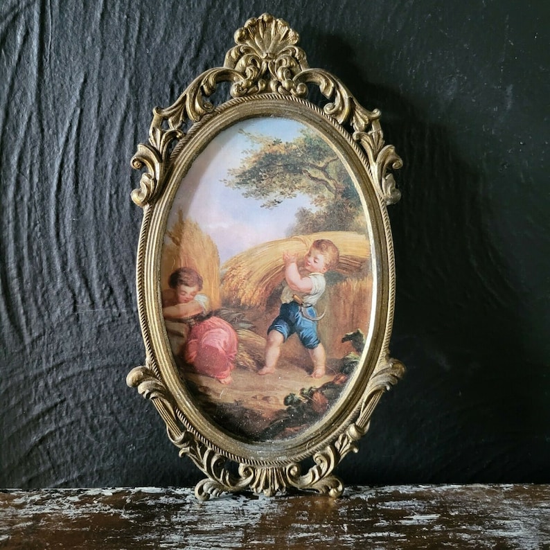 May include: A gold-toned ornate oval picture frame with a detailed design. The frame holds a vintage painting of two children in a field of wheat.