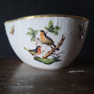May include: A white porcelain bowl featuring a textured basket-weave pattern on the exterior. The bowl is decorated with hand-painted illustrations of two birds perched on a branch with green leaves and a small butterfly on the side, finished with a gold rim.
