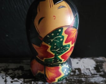 May include: A wooden Kokeshi doll featuring a smooth, rounded head with painted black hair and closed eyes. The body is decorated with a vibrant pattern of red and orange maple leaves against a dark green background with gold accents.