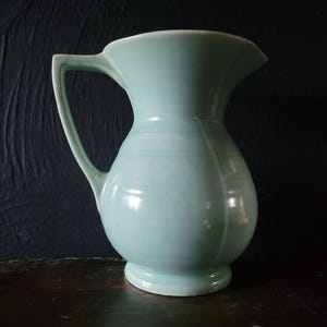 Puede incluir: Una jarra de cerámica verde claro con una boca ancha y un cuerpo redondeado. La jarra tiene un acabado liso y brillante y un labio ligeramente acampanado.