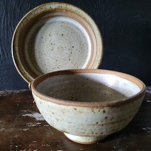 Op de afbeelding: Een witte keramische kom met een bruine rand en gespikkelde glazuur. De kom staat op een houten oppervlak. Een bijpassend wit keramisch bord met een bruine rand en gespikkelde glazuur is op de achtergrond.