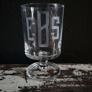 May include: A clear glass goblet with a wide bowl and a short stem. The letters "ERS" are etched into the glass in a decorative font. The goblet is sitting on a distressed wooden surface.