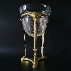 Puede incluir: Un centro de mesa antiguo de cristal y latón. El cuenco de cristal transparente tiene una textura agrietada y se asienta en un soporte de latón decorativo. El soporte presenta detalles ornamentados y una base de tres patas, creando una elegante exhibición.
