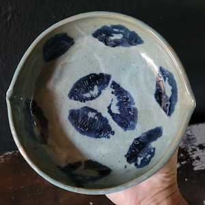 May include: A light blue ceramic bowl with a lip on one side. The bowl is decorated with a pattern of blue lip prints.