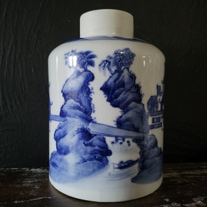 Op de afbeelding: Een witte keramische pot met een blauw en wit geschilderd ontwerp van een landschap met bergen, bomen en een brug. De pot heeft een rond lichaam en een brede opening.