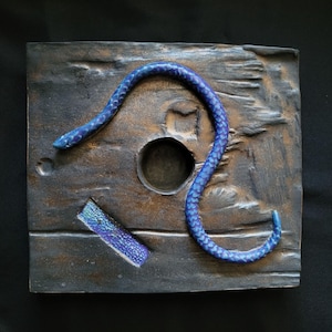 May include: A square, textured, wooden-like sculpture with a blue snake coiled around a circular hole. A broken piece of the same material is near the hole. The snake has a scale pattern and is a vibrant blue color.