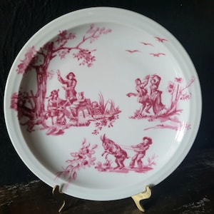 May include: A white porcelain plate with a pink floral and pastoral scene. The scene depicts figures in a wooded setting with a goat and a tree.