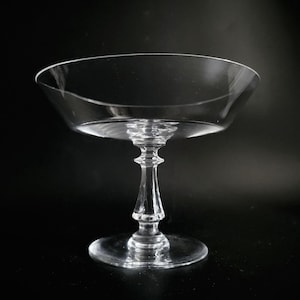 Könnte beinhalten: Ein klares Champagner-Coupé-Glas mit einer breiten, flachen Schale und einem dekorativen Stiel. Das Glas steht auf einem runden Fuß und vor einem dunklen Hintergrund. Das Glas besteht aus klarem Glas.