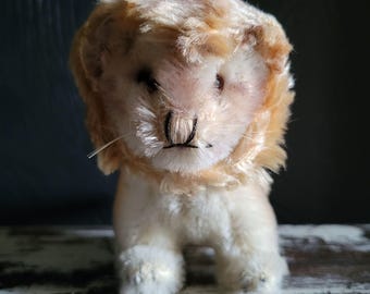 Puede incluir: Un juguete de peluche vintage con forma de león, con cuerpo color crema y melena marrón claro. El león tiene ojos pequeños y oscuros, nariz cosida en negro y rostro detallado. El juguete es de material suave y afelpado, en posición sentada.