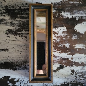May include: A rectangular, vintage mirror with a gold-colored frame and a black inner frame. The mirror is long and narrow, reflecting a partial view of a room. The background is a distressed, white-painted wooden surface.