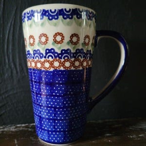 Könnte beinhalten: Eine hohe blaue und weiße Keramiktasse mit einem braunen und grünen Blumenmuster. Die Tasse hat einen großen Henkel und einen breiten Boden.