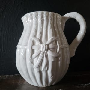 May include: White ceramic pitcher with a textured, woven design and a large bow on the front. The pitcher has a handle and a wide mouth.