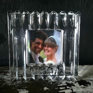 May include: A clear, heavy glass picture frame featuring a thick, vertical, faceted, and ribbed border design. The frame holds a square photograph of a smiling couple, with one person wearing a white veil, displayed in the center of the glass structure.