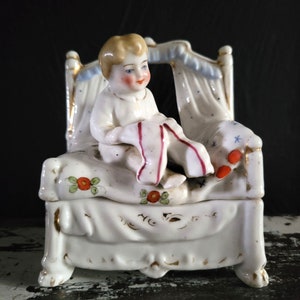 May include: A white porcelain figurine of a baby sitting up in a bed. The bed has a canopy and is decorated with gold trim, red and green floral accents, and blue accents. The baby is holding a red and white garment.