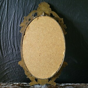 May include: An oval-shaped brass picture frame with an ornate design. The frame is empty and has a brown cardboard backing.