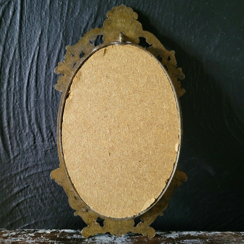 May include: An oval-shaped brass picture frame with an ornate design. The frame is empty and has a brown cardboard backing.