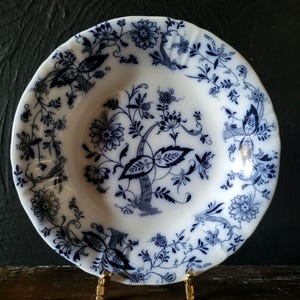 May include: White ceramic plate with a blue floral pattern. The plate features a central tree-like design and a border of flowers and leaves. The plate is displayed on a gold-coloured stand.