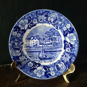 May include: Blue and white porcelain plate with a floral border and a scene of a village by a river with a sailboat.