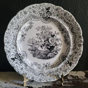 May include: A white ceramic plate with a black floral and landscape design. The plate has a scalloped edge and features a central image of a vase of flowers and a ship. Decorative tableware.