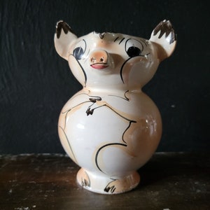 May include: A white ceramic piggy bank with brown and black painted details. The pig has large eyes and a pink snout. The piggy bank is sitting on a wooden surface.