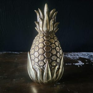 Puede incluir: Una escultura de piña de latón macizo con un cuerpo de textura en forma de diamante y hojas lisas y puntiagudas en la parte superior e inferior. La superficie metálica muestra una pátina de color marrón dorado con reflejos brillantes.