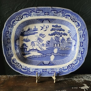 May include: An oval ceramic platter featuring a blue and white transferware pattern. The design depicts a landscape with a pagoda, a bridge, a willow tree, and two birds in flight. The wide border displays a geometric and floral motif.