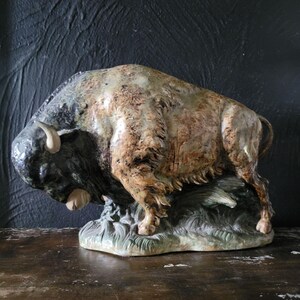 May include: A ceramic sculpture of a brown and black bison with a textured surface. The bison is standing on a green base with a textured surface.