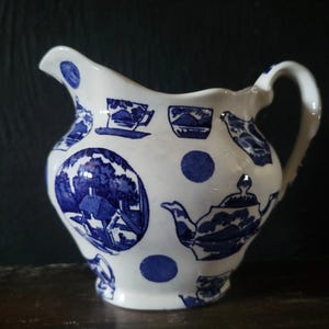 Puede incluir: Una jarra de crema de cerámica blanca con un patrón azul y blanco de tazas de té, teteras y una escena de paisaje. La jarra tiene un cuerpo redondeado y un asa.