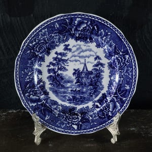May include: A round ceramic plate featuring a blue and white transferware pattern. The center depicts a landscape with a tower, trees, and a small boat on water. The border is decorated with large floral blooms and a scalloped edge design.