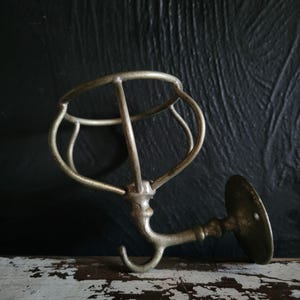 May include: Antique brass-toned wall-mounted soap dish holder with a circular cage design and a curved hook. The holder has a round base with a single screw hole for mounting. The metal has a weathered appearance.