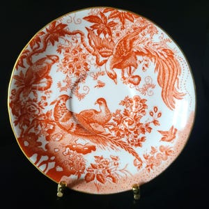 May include: White ceramic plate with a detailed orange bird and floral design. The plate features a gold rim and a decorative pattern of birds, flowers, and foliage. The design is a classic, elegant style.
