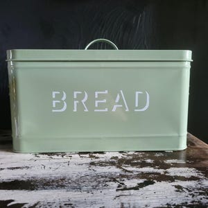 May include: A rectangular, light green bread box with a curved handle on top. The word "BREAD" is printed in white, block letters on the front. The bread box is made of metal and sits on a wooden surface.