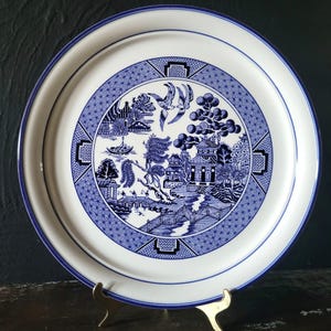 May include: A white and blue decorative plate with a classic Willow pattern design. The plate features a blue border and a detailed landscape scene with birds, bridges, and buildings. The plate is displayed on a gold-colored stand.