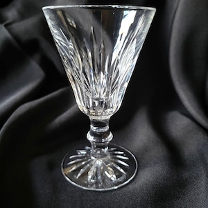 May include: A clear crystal wine glass with a detailed, etched pattern. The glass has a wide, conical bowl, a slender stem, and a circular base. The etched design features vertical lines and a starburst pattern on the base.