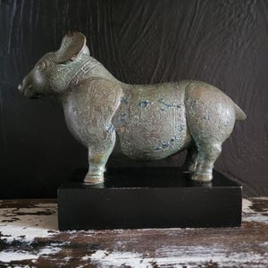 Op de afbeelding: Een bronzen varkenssculptuur met ingewikkelde geëtste patronen, geplaatst op een zwarte rechthoekige basis. Het varken heeft een verweerde uitstraling. De donkere achtergrond accentueert de vorm en details van de sculptuur.