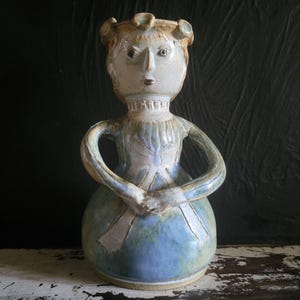 Puede incluir: Figura de cerámica de una mujer con un vestido azul y los brazos cruzados. La figura tiene la cara y el pelo de color marrón claro con detalles en azul y verde. El vestido es azul claro con una falda azul más oscura.