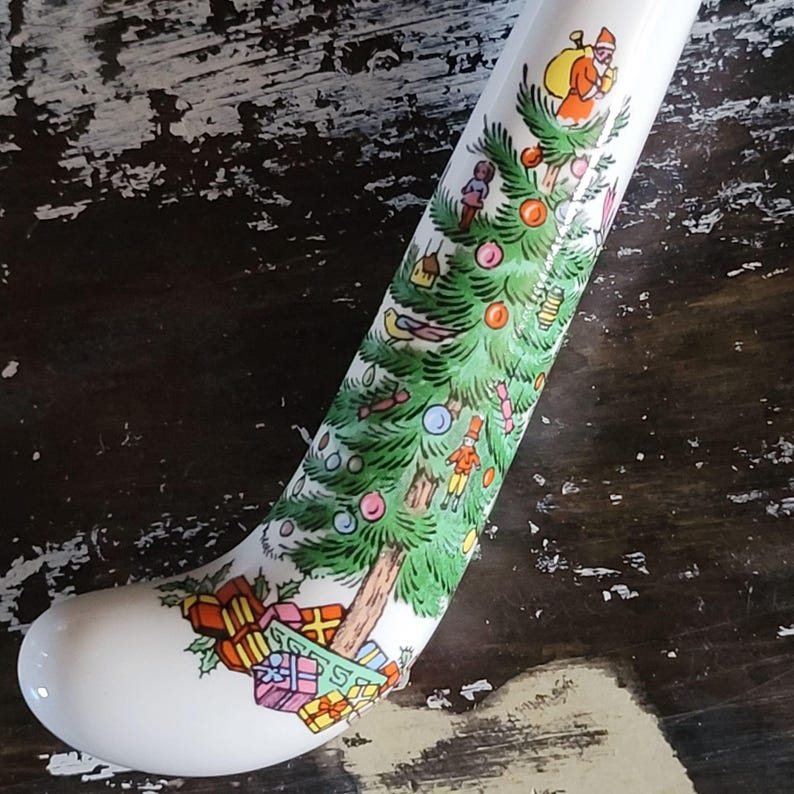 May include: A white ceramic Christmas stocking with a festive design. The stocking features a green Christmas tree adorned with ornaments, Father Christmas, and wrapped gifts. The design is in a colorful, cartoon style.