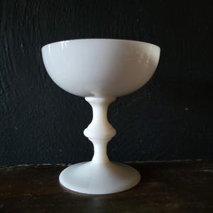 May include: A white, stemmed coupe glass with a rounded bowl and a pedestal base. The glass has a classic design, with a wide, shallow bowl and a slender stem. The glass is made of a smooth, opaque material.