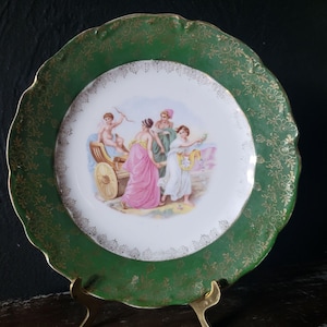May include: A decorative plate with a green border and gold floral accents. The center features a classical scene with figures in flowing robes and a chariot. The plate is displayed on a gold stand.