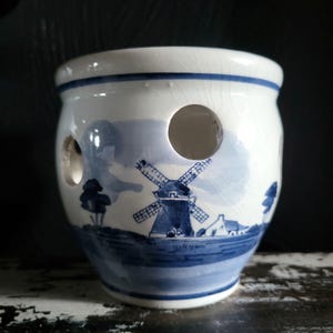 May include: A white ceramic garlic keeper with a blue and white windmill design. The keeper has three circular holes and a blue rim. The design features a windmill, trees, and a house, evoking a Dutch landscape.