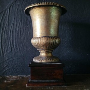 May include: A gold-colored metal urn with a fluted design sits on a dark wood base. The urn is on a dark wood surface.