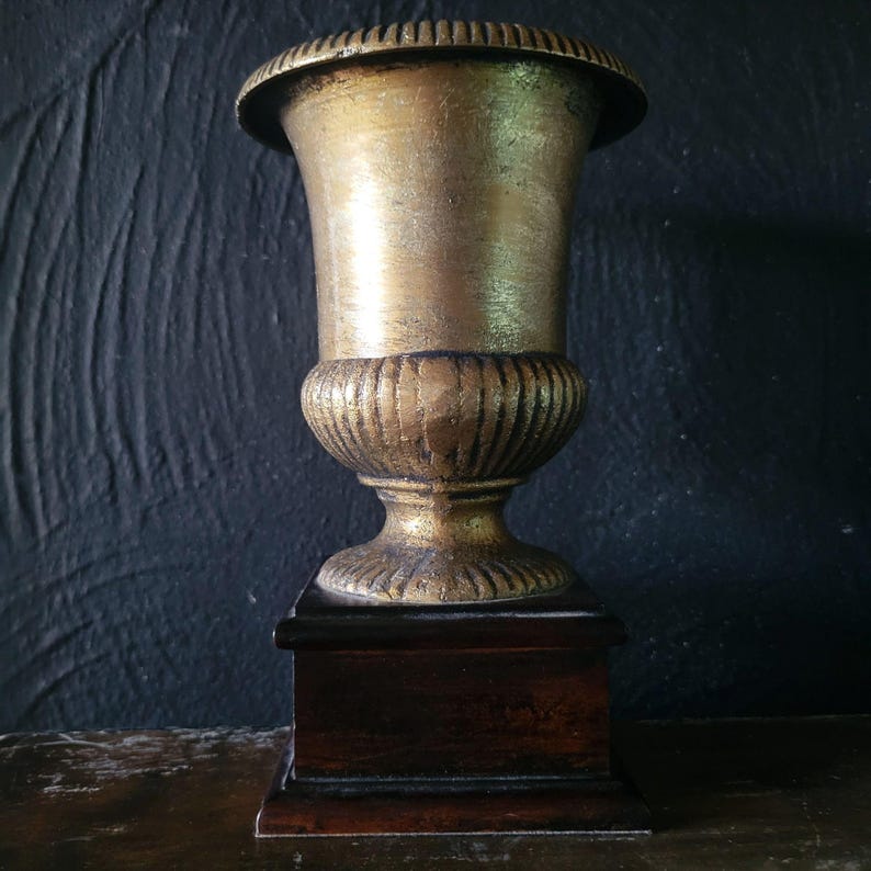 May include: A gold-colored metal urn with a fluted design sits on a dark wood base. The urn is on a dark wood surface.