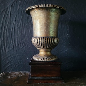 May include: A gold-colored metal urn with a fluted base and a square wooden base. The urn is on a dark wooden surface.