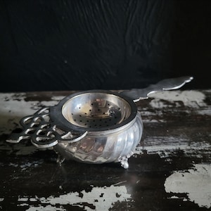 Puede incluir: Un colador de té de metal plateado con un cuenco de base perforada, un mango decorativo calado en un lado y un mango largo y plano en el lado opuesto. El colador se apoya sobre pequeñas patas ornamentadas y tiene un cuerpo redondeado y estriado.